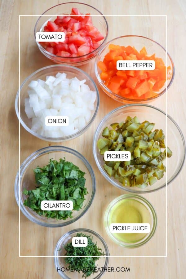 Seven glass bowls on a wooden surface contain labeled ingredients: chopped tomato, bell pepper, onion, pickles, cilantro, dill, and a small amount of pickle juice.