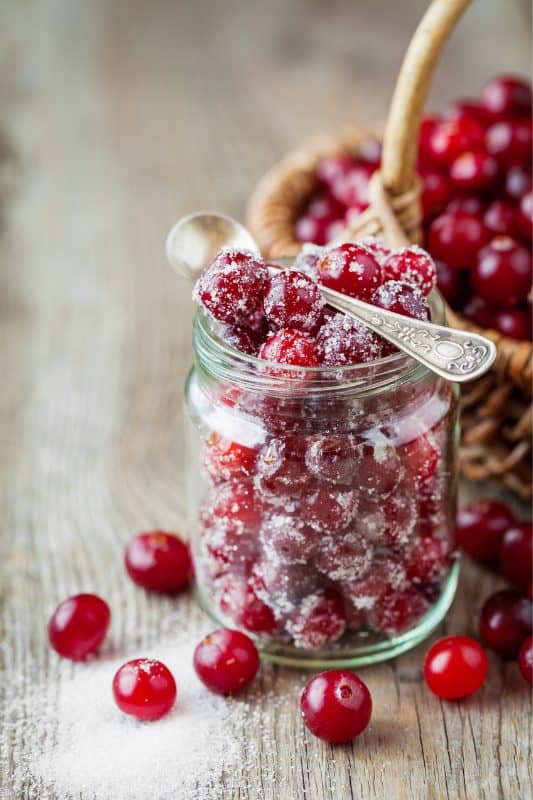 A glass jar filled with sugared cranberries sits on a wooden surface, with a vintage spoon inside. Loose cranberries and a basket of cranberries are in the background.