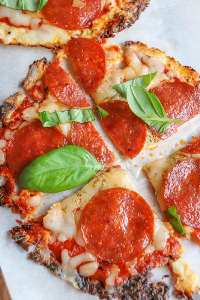 A close-up of a sliced pepperoni pizza with melted cheese, tomato sauce, and fresh basil leaves on a crispy, golden-brown crust, served on parchment paper.