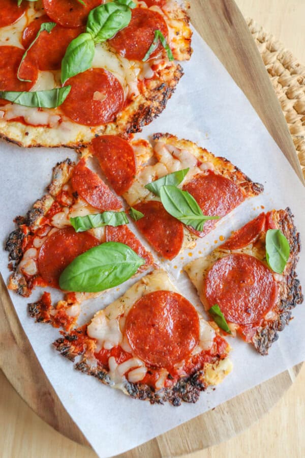 Pepperoni pizza sliced on parchment paper, topped with melted cheese, pepperoni, and fresh basil leaves, served on a wooden board.