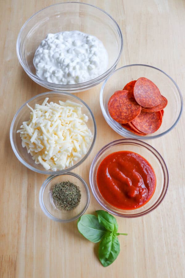 Five glass bowls on a wooden surface containing cottage cheese, pepperoni slices, shredded cheese, pizza sauce, and Italian seasoning, with a fresh basil leaf placed beside them.