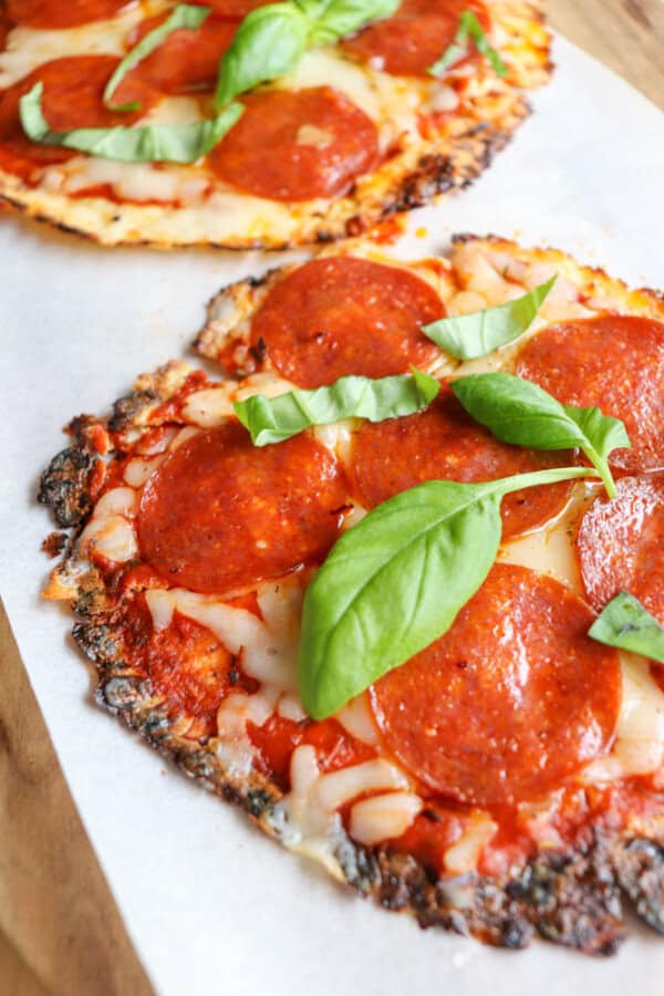 Two pepperoni pizzas with melted cheese and tomato sauce on a crispy crust, each topped with fresh basil leaves, served on white parchment paper.