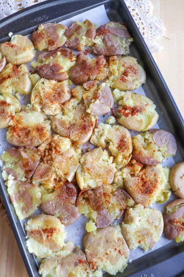 A baking tray lined with parchment paper holds smashed, roasted potatoes sprinkled with spices, ready to serve. Some potato skins are crispy and browned, and a lace cloth is partially visible in the background.