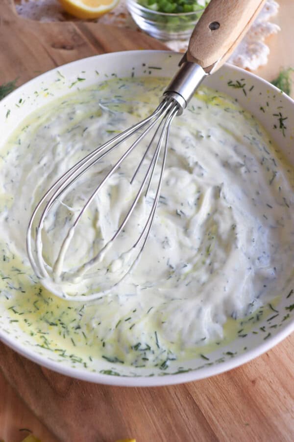 A metal whisk rests in a white bowl filled with creamy dill sauce or dip, with green herb flecks visible throughout the mixture. The bowl sits on a wooden surface.