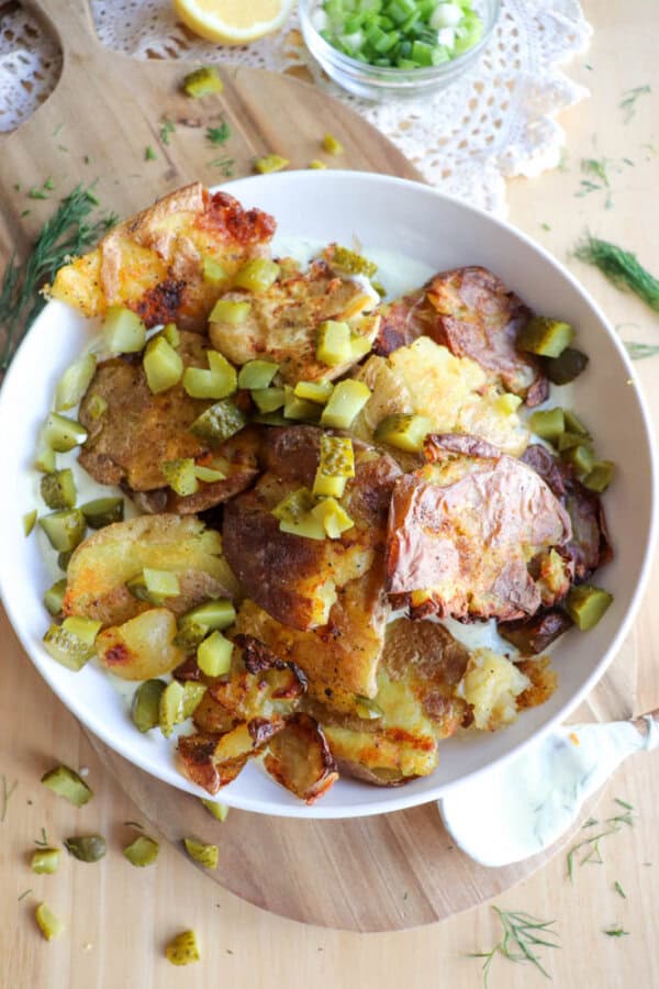 A white bowl filled with crispy smashed potatoes topped with chopped pickles, set on a wooden board. Fresh dill, green onions, and a small bowl of white sauce are nearby.