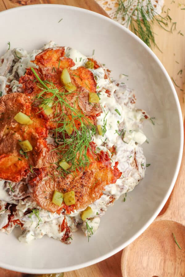 A white bowl filled with crispy fried potato cakes topped with chopped pickles and fresh dill, served over a creamy salad mixture with herbs on a wooden surface.