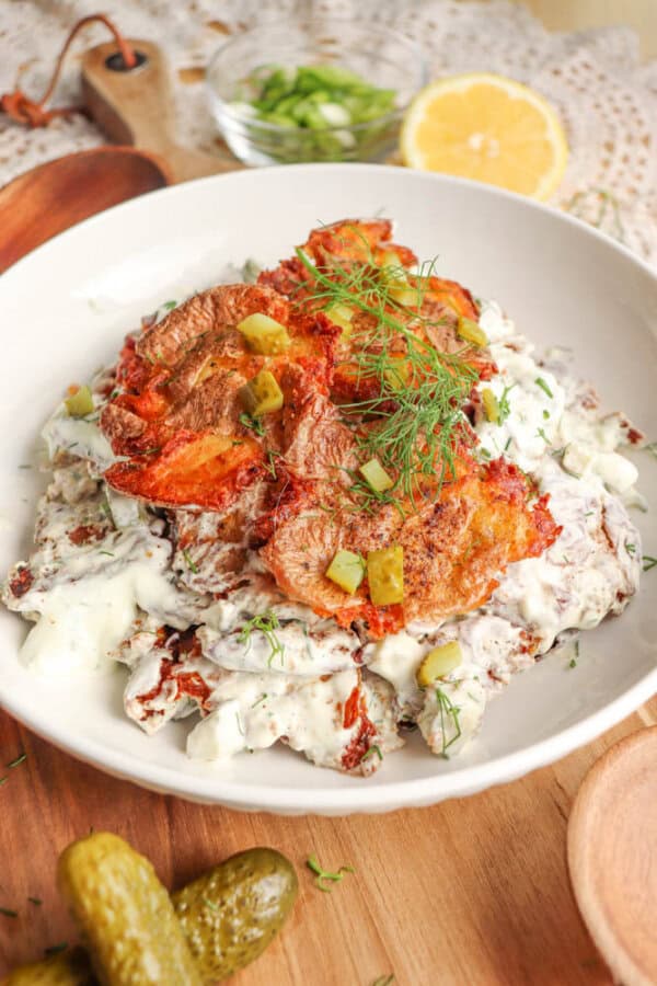 A white plate holds crispy pieces of fried fish on creamy, herbed sauce with chopped pickles and fresh dill, served on a wooden board with sliced pickles, lemon, and green garnish in the background.