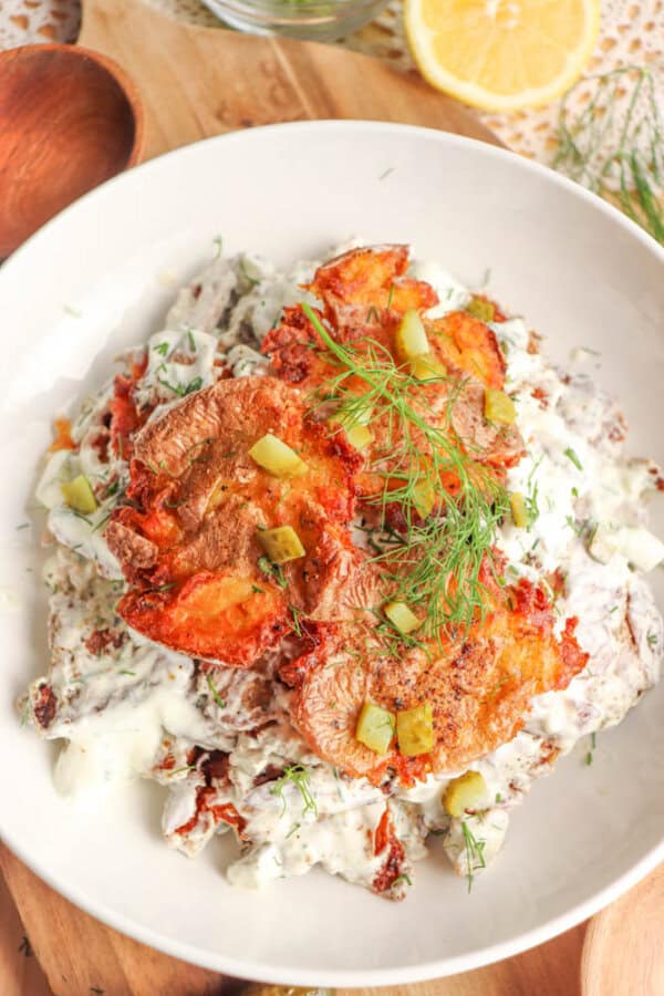 A white bowl filled with creamy salad topped with golden, crispy fried tomatoes, diced pickles, and fresh dill, set on a wooden surface with a lemon slice and dill fronds nearby.