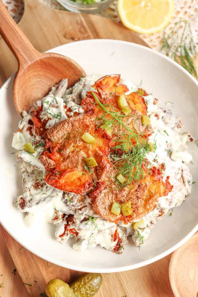 A white bowl filled with a creamy salad topped with golden, crispy roasted slices, fresh dill, and diced pickles. A wooden spoon rests in the bowl, with part of a lemon and pickles visible in the background.
