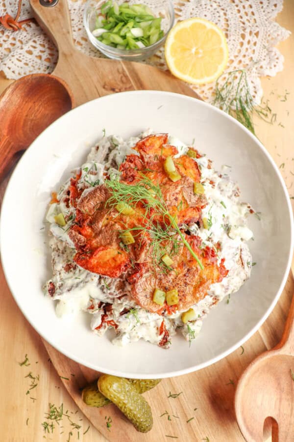 A white bowl filled with potato pancakes topped with creamy sauce, fresh dill, and chopped pickles. Sliced lemon, green onions, wooden utensils, and a lace doily are on the wooden table around the bowl.