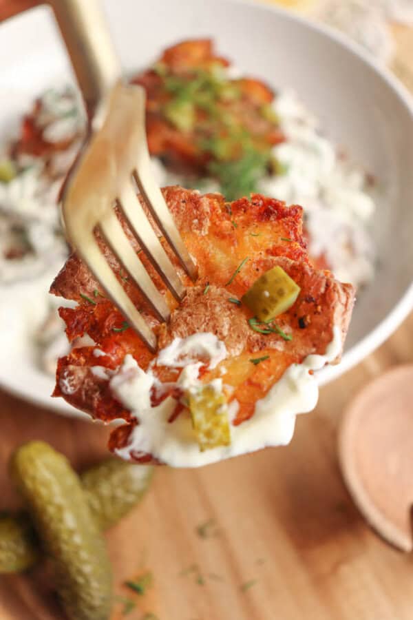 A close-up of a golden-brown smashed potato on a fork, topped with diced pickles and drizzled with creamy white sauce. More potatoes and pickles are visible on a plate and wooden surface in the background.