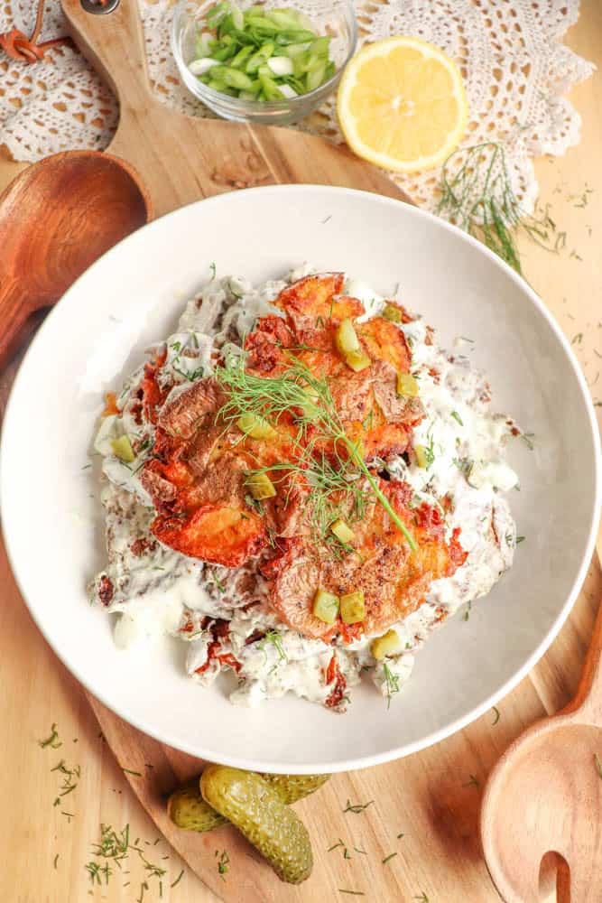 A white bowl filled with potato pancakes topped with creamy sauce, fresh dill, and chopped pickles. Sliced lemon, green onions, wooden utensils, and a lace doily are on the wooden table around the bowl.