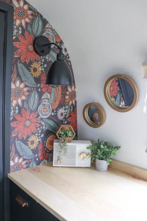 A small desk with a light wood surface sits against a floral-patterned wall, evoking the charm of an argosy airstream restoration. Two round mirrors, a black wall lamp, vintage radio, potted succulent, and green plant complete the look.