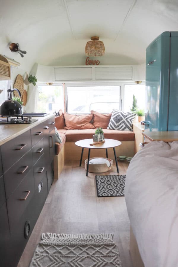 Cozy, modern Argosy Airstream restoration with a kitchen on the left, blue fridge on the right, and a seating nook with pink cushions and throw pillows by the back windows. A small round coffee table and decor complete the space.