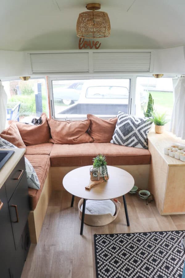 A cozy Argosy Airstream restoration features a brown L-shaped sofa, patterned pillows, round wooden coffee table, rug, plants, and large rear window. Natural light fills the space, creating a warm, inviting atmosphere.