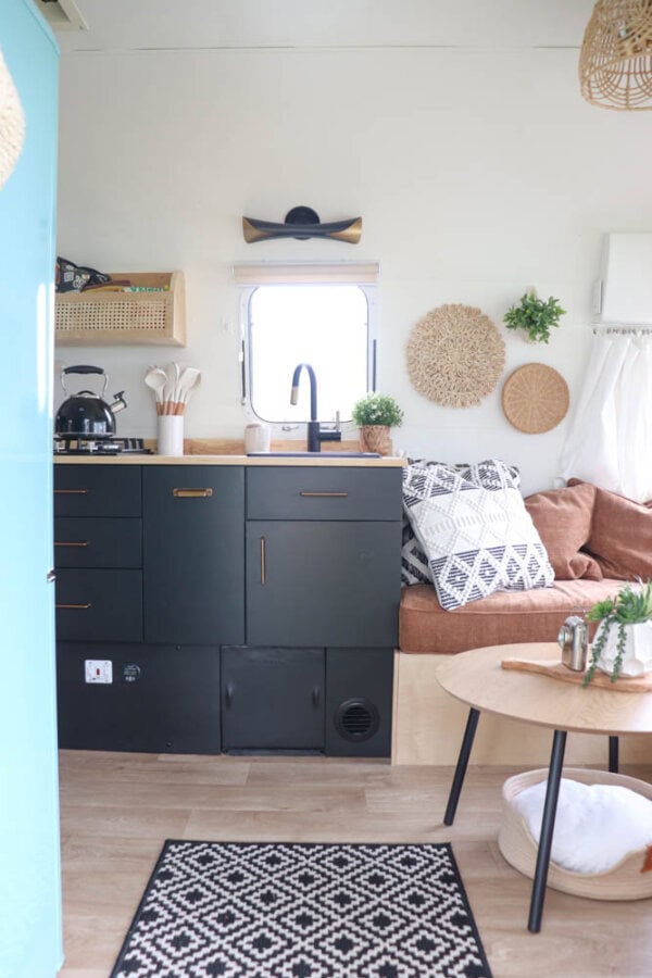 Modern Argosy Airstream restoration with black cabinets, tan sofa with patterned pillows, small round table, potted plants, woven wall decor, a tea kettle, and a window above the sink letting in natural light.