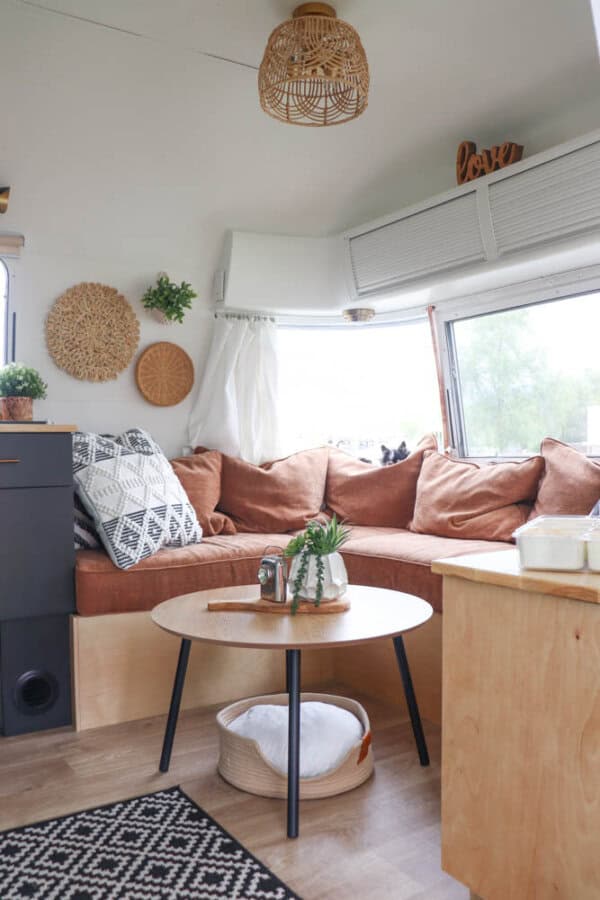 Cozy Argosy Airstream restoration interior with a brown L-shaped sofa, geometric cushions, round wooden coffee table with plants, black-and-white rug, wicker wall decor, and large windows letting in natural light.
