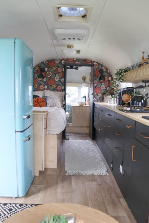 A cozy Argosy Airstream restoration featuring wood floors, black lower cabinets, wood countertops, a blue retro fridge, and a rear bed. Colorful floral wallpaper brightens the walls above the bed, with plants and kitchen items on the counters.