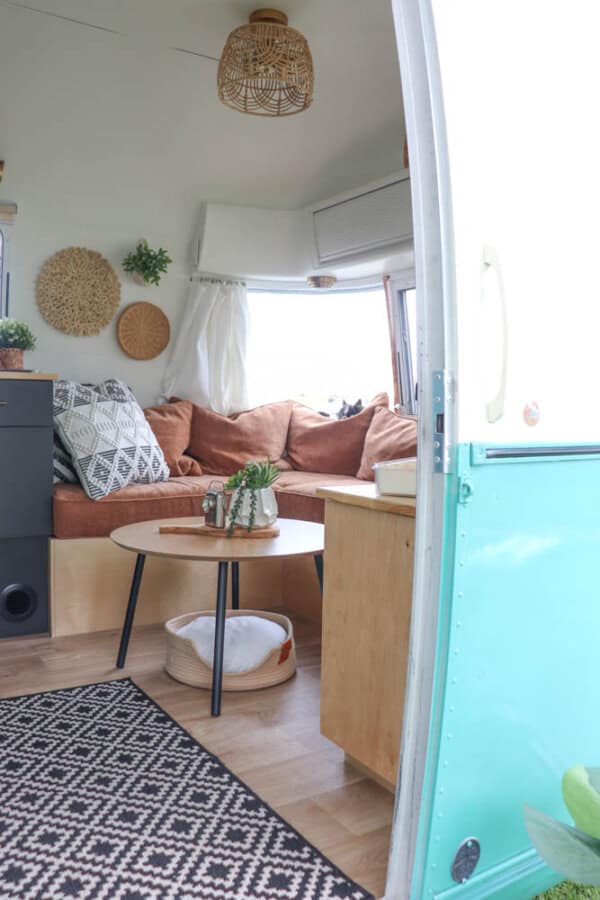 A cozy camper interior in an Argosy Airstream restoration with a brown sectional sofa, geometric pillows, a round wooden coffee table, woven wall decor, and a black-and-white patterned rug. Light streams through a window with white curtains.
