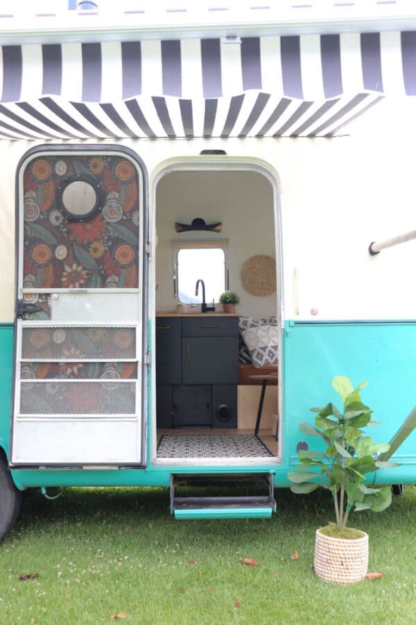 A turquoise and white Argosy Airstream restoration features an open door revealing a small, modern kitchen. A black and white striped awning extends above, while a potted plant sits on the grass outside the steps.