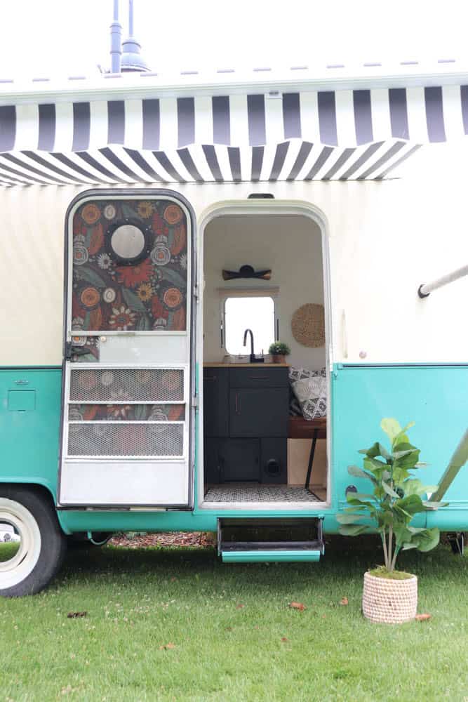 A turquoise and white Argosy Airstream camper van with a black-and-white striped awning. The door is open, revealing a cozy, restored interior with cabinets, a window, and a potted plant on the grass outside.