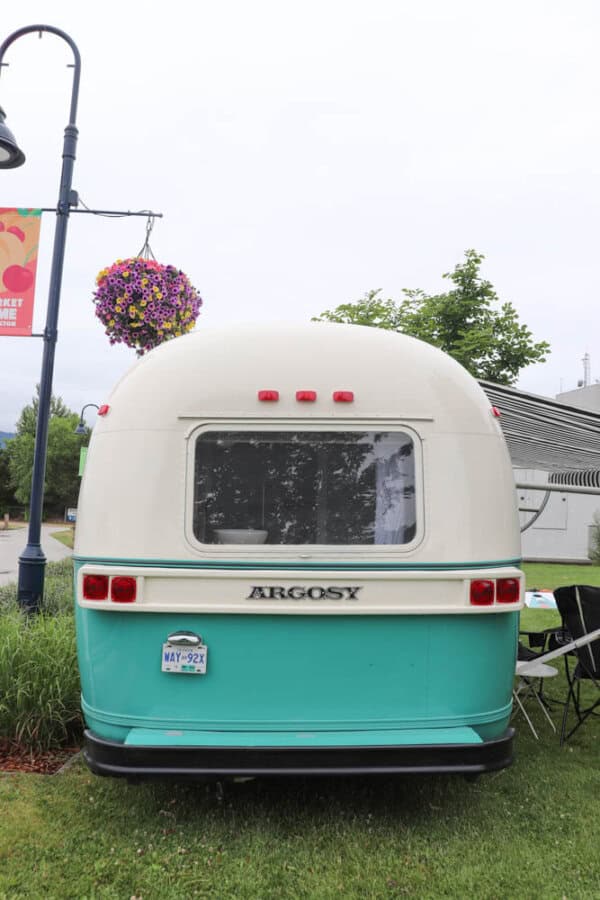 A vintage turquoise and white Argosy camper, recently enjoying an argosy airstream restoration, is parked on grass near a street lamp with a hanging basket of purple flowers. Trees and a foldable table and chair sit beside the camper.