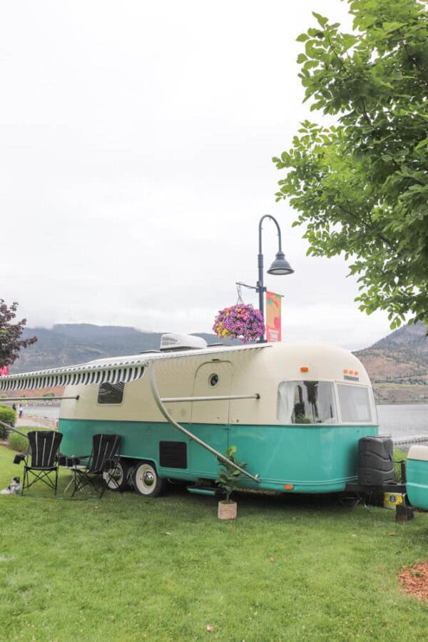 A vintage turquoise and cream Argosy Airstream camper trailer with an extended awning is parked on grass near a lake. Black chairs sit outside, and flowers hang from a nearby lamp post, with mountains visible in the background, showcasing careful restoration.