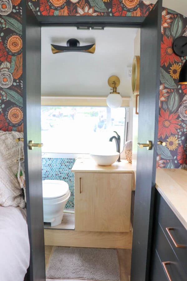 A small bathroom in an argosy airstream restoration features colorful floral wallpaper, a white toilet, a light wood vanity with a round vessel sink, modern faucet, and window. Part of a bed and rug are visible in the foreground.