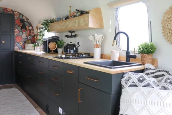 A modern kitchen inside an Argosy Airstream restoration features dark cabinets with gold handles, a wooden countertop, a black sink, gas stove, potted plants, open shelving, and patterned decor accents. Natural light streams in through a window.