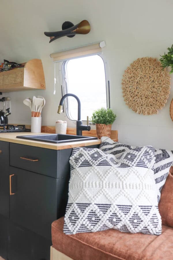 A cozy camper kitchen in an Argosy Airstream restoration features a black sink, gold faucet, wooden counter, utensils in a holder, potted plants, and a couch with a patterned pillow. Woven wall decor and a window brighten the space.
