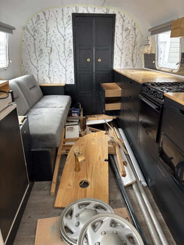 An Argosy Airstream restoration in progress: an RV interior features a gray sofa, black cabinets, light wood countertops, open drawers, scattered wooden panels, two metal hubcaps, tools on the floor, and a back wall with white tree-patterned wallpaper.