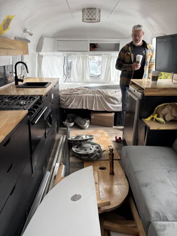 A man holding a cup stands inside a renovated Argosy Airstream camper van with wooden countertops, a stove, a bed in the back, and tools and materials on the floor, hinting at ongoing restoration work.