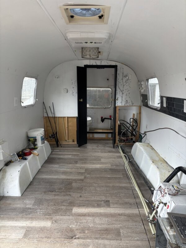 The interior of an empty, partially renovated Argosy Airstream shows exposed walls, wood-look flooring, plumbing pipes, tools, and a view into a small bathroom at the rearโperfect for your restoration project.