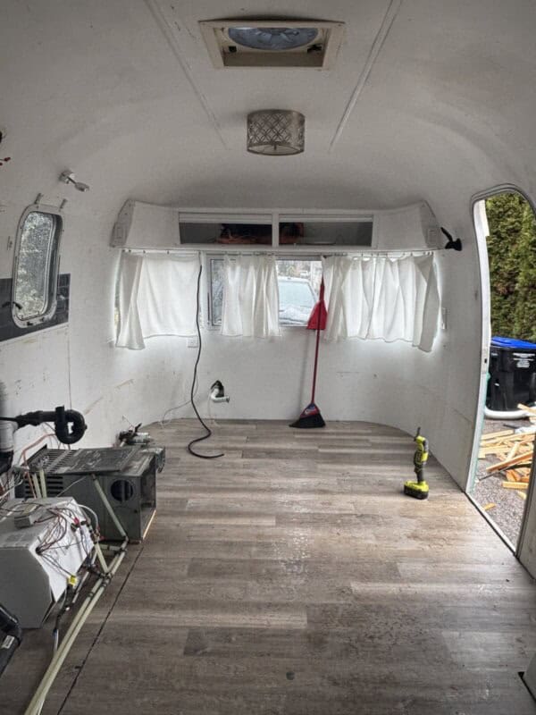 The interior of an empty Argosy Airstream mid-restoration features wood flooring, white walls, and a small window with white curtains. Tools and equipment lie on the floor, while exposed wires run along the left side.
