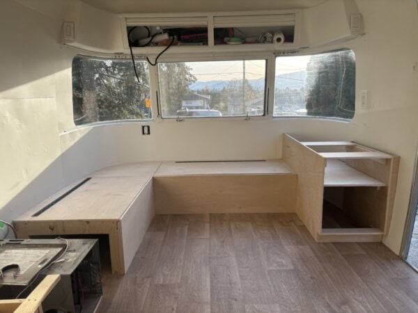 A partially renovated Argosy Airstream interior features unfinished wooden bench seating, open shelves, and large windows letting in natural lightโa promising restoration project. The floor is covered with wood-look vinyl planks and tools are visible in the foreground.