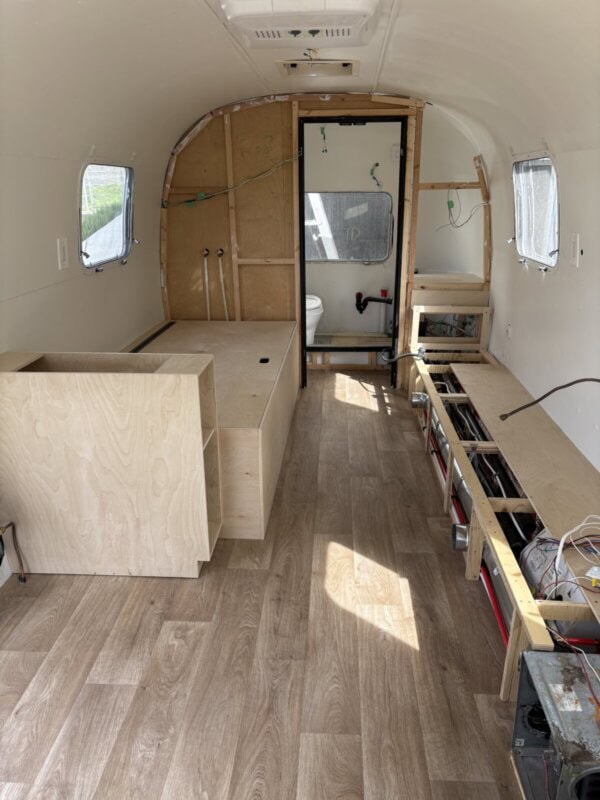 Interior of an Argosy Airstream under restoration, featuring wooden flooring, an unfinished bed frame, exposed wiring, and a visible bathroom at the back. The space is bright with natural light streaming through the windows.