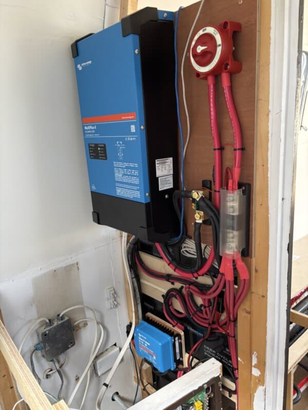 A blue Victron Multiplus-II inverter is mounted on a wooden wall in this argosy airstream restoration, with thick red and black cables, a large red battery disconnect switch, fuse, and other electrical components neatly installed nearby.