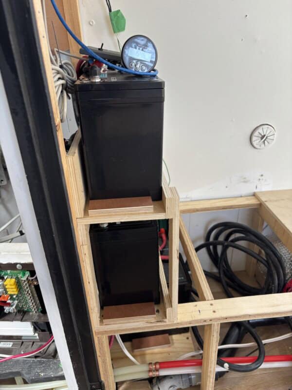 Two black batteries are stacked vertically on wooden supports with a small digital meter on topโtypical of an Argosy Airstream restoration. Wires, cables, and additional electronics with coiled cords are visible nearby.