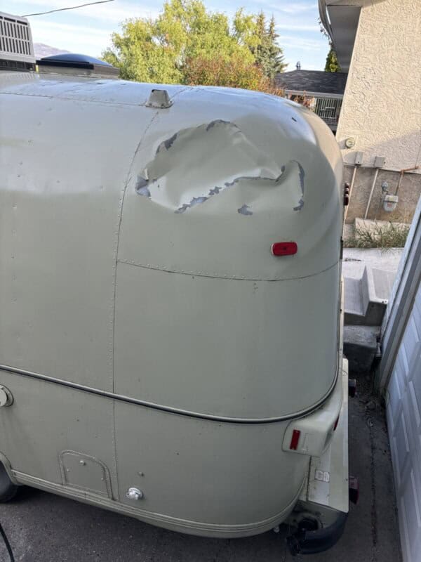 A beige Argosy Airstream trailer with a large area of paint peeling off near the top rear corner, exposing the underlying surfaceโperfect for a restoration project. The trailer is parked near a garage on a concrete driveway.