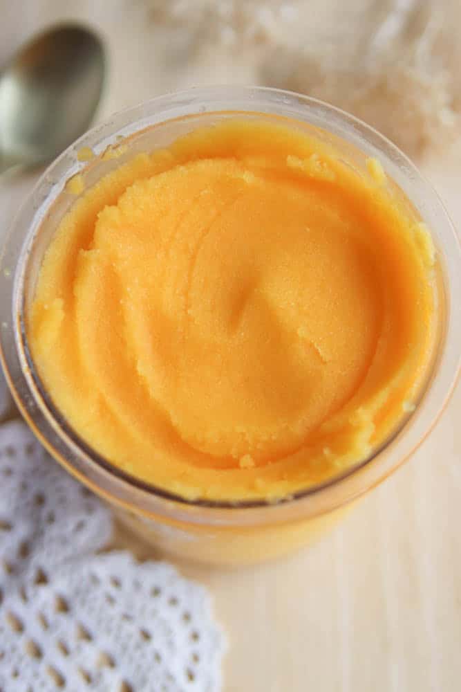 A glass jar filled with smooth, bright orange mango sorbet sits on a light surface next to a gold spoon and a white crocheted cloth.