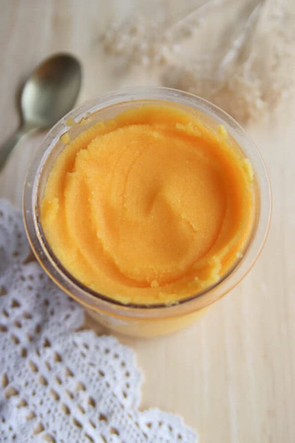 A glass jar filled with smooth, bright orange sorbet sits on a wooden surface next to a silver spoon and a white crocheted cloth. Delicate dried flowers are visible in the background.