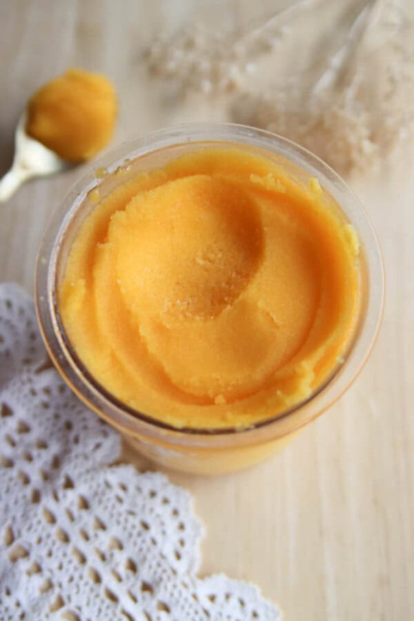 A glass jar filled with smooth, orange mango sorbet sits on a light wooden surface next to a white crochet cloth. A gold spoon with a scoop of sorbet is in the background.