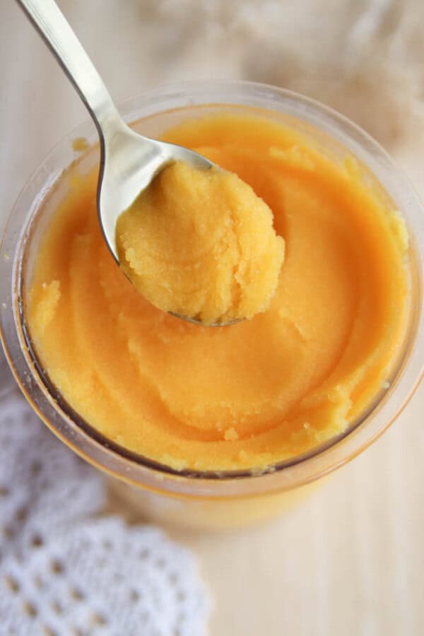 A close-up of a glass jar filled with smooth, yellow mango sorbet. A spoon is holding a scoop above the jar. A white textured cloth is partially visible beside the jar on a wooden surface.