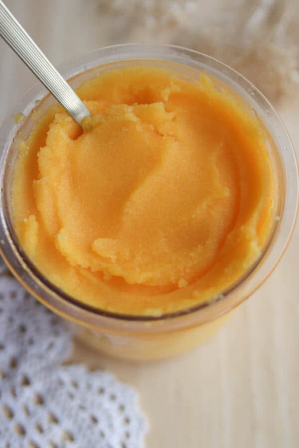 A glass jar filled with creamy, orange-colored mango sorbet, with a metal spoon dipped into it, sits on a wooden surface next to a white crocheted cloth.