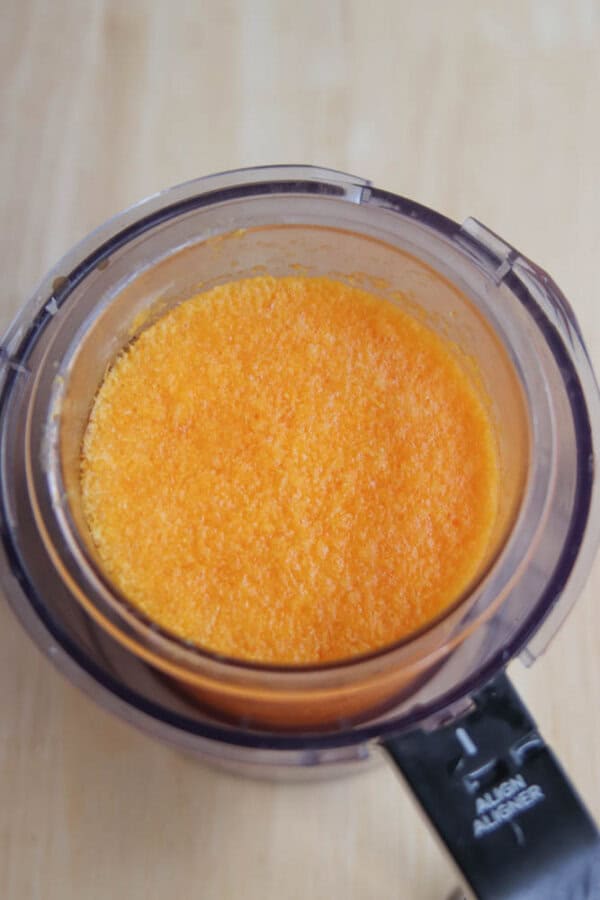 A food processor bowl filled with finely blended or grated orange carrots, viewed from above on a light wooden surface.