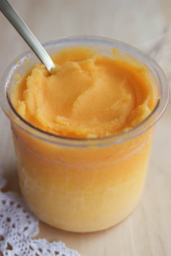 A glass jar filled with smooth, orange-colored sorbet or frozen dessert, with a spoon inside. The jar sits on a light wooden surface next to a piece of lace fabric.