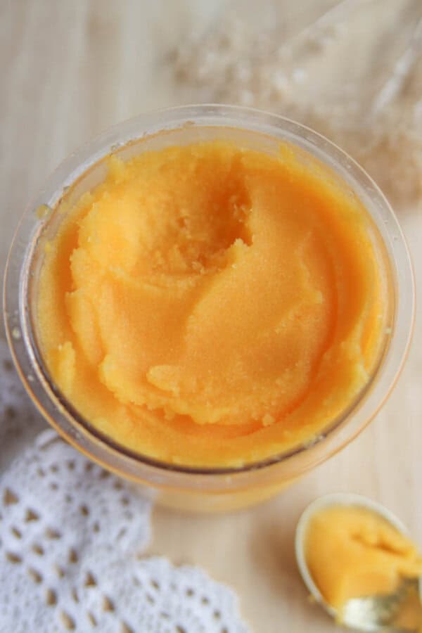 A glass jar filled with smooth, yellow-orange mango sorbet sits on a light wooden surface next to a lace doily and a spoon holding a small scoop of the sorbet.