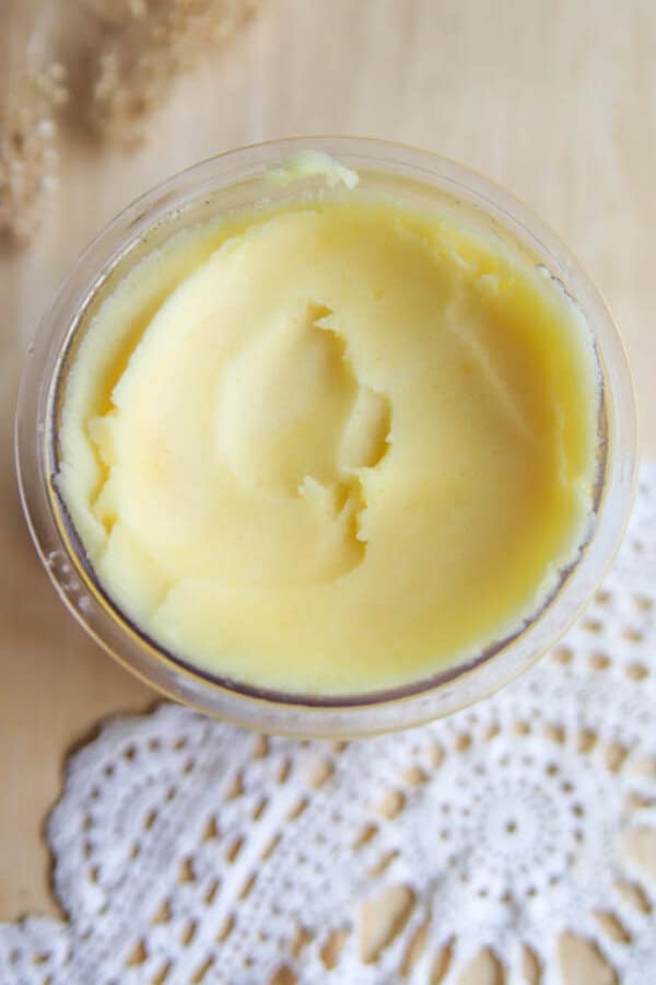 A glass bowl filled with creamy, yellow homemade body butter sits on a wooden surface next to a white lace doily. The whipped, smooth texture is reminiscent of ninja creami peach sorbet.
