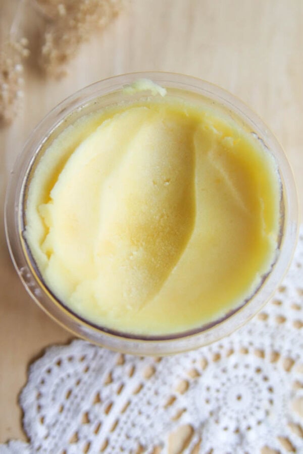A clear container filled with smooth, yellow butter sits on a wooden surface next to a white lace doily, reminiscent of the creamy texture found in Ninja Creami peach sorbet.