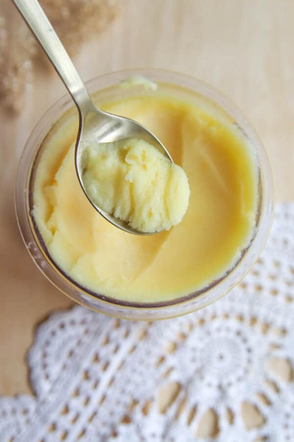 A spoon scoops creamy yellow ghee from a small glass jar, set on a wooden surface next to a white crocheted doily—perfect beside a bowl of homemade Ninja Creami peach sorbet.
